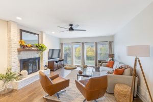 A cozy living room with white walls, wood flooring, and large windows with beige curtains. The room features a white painted brick fireplace, a ceiling fan, a gray sofa with orange pillows, two brown leather chairs, a round glass coffee table, and a TV on a wooden stand.