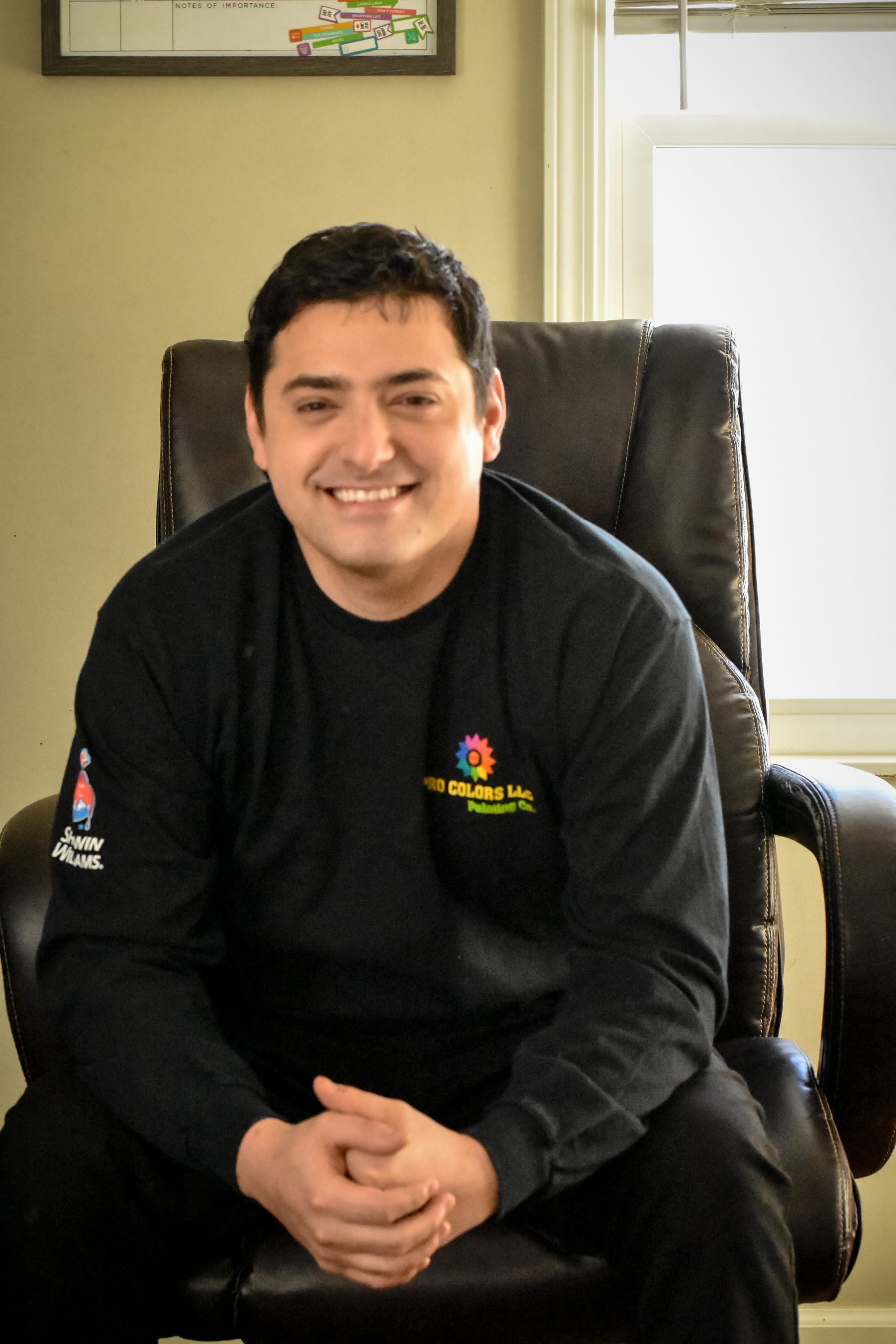 A person with short dark hair is sitting and smiling. They are wearing a dark long-sleeve shirt with a colorful logo on the chest and left sleeve. The background features part of a window and a wall with a framed item on it.