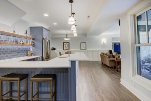 A spacious, modern kitchen and living area with white walls and light hardwood floors. The kitchen features a gray island with two barstools, pendant lights, and a tiled backsplash. In the background, there's a cozy seating area with a sofa and a TV.