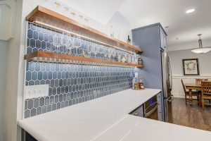 Modern kitchen with a white marble countertop and hexagonal tiled backsplash in shades of blue. Above the counter are two wooden shelves holding glassware and bottles. The kitchen fridge, dining table, and chairs are visible in the background.