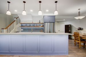 A spacious kitchen featuring a large island with a light blue panel design and a white countertop. Hanging pendant lights illuminate the area. Behind the island, there's a blue-tiled backsplash, open shelves, and a stainless steel refrigerator. On the right, a wooden dining table with chairs is visible.