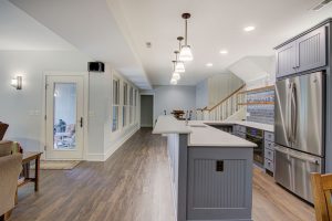 Spacious kitchen with long island countertop, modern pendant lights, stainless steel appliances, and gray cabinets. Hardwood floors extend through the open layout leading to a staircase. Sliding glass door on the left opens to a brightly lit room.