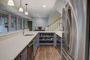 A modern kitchen featuring a large island with a sink and marble countertop, pendant lights above, and stainless steel appliances. Cabinets and drawers are open, revealing dishes and glasses. To the side, there is a dining area and a staircase ascending to the upper floor.